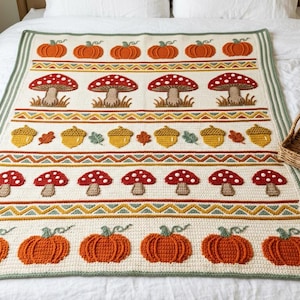 May include: A rectangular crochet blanket featuring a cream-colored base with horizontal rows of orange pumpkins, red and white mushrooms, and yellow acorns. The design includes geometric zigzag borders in green, yellow, and orange tones.