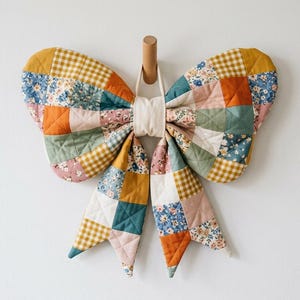 May include: A large, quilted fabric bow shaped like a butterfly, constructed from a patchwork of squares featuring floral patterns, solid mustard yellow, sage green, and white gingham fabrics, with a central cream-colored fabric knot.