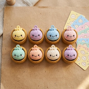 May include: Eight small, round, crocheted plush toys in various pastel colors, each featuring a smiling face with black eyes and pink cheeks. Each toy sits inside a small, brown, crocheted bowl-like base. Several illustrated cards with dumpling graphics are stacked to the right.
