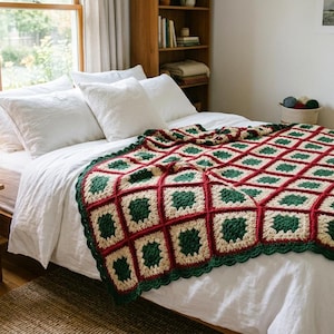 May include: A handmade crochet blanket featuring a repeating granny square pattern in deep forest green, cream, and dark red yarn. The blanket is draped over a bed with white bedding, showcasing a scalloped edge border and a textured yarn finish.
