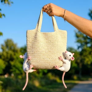 May include: A cream-colored crocheted tote bag featuring two small, white, handmade crocheted rat figures attached to the lower sides. The bag has two sturdy handles and a textured, woven appearance, with the rat figures showing pink ears.