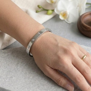 Op de afbeelding: Een zilverkleurige metalen armband met een reeks ingelegde heldere stenen en gegraveerde schroefkopmotieven langs de buitenste band. De armband heeft een gepolijste afwerking en een scharnierend ontwerp, gedragen met een gouden ring.