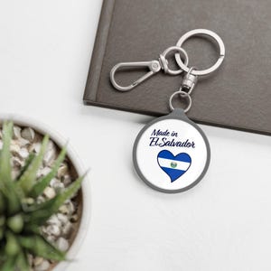 Puede incluir: Un llavero circular con fondo blanco que presenta el texto Made in El Salvador en letra cursiva azul oscuro sobre un gráfico en forma de corazón que contiene las franjas horizontales azules y blancas y el emblema de la bandera salvadoreña.
