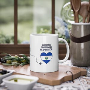 May include: A white ceramic mug featuring the text MI CORAZÓN TIENE FORMA DE EL SALVADOR in dark blue letters above a heart-shaped graphic containing the El Salvador flag, with a small black dashed line and airplane icon trailing to the left.