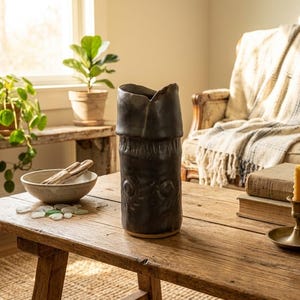 May include: A tall, cylindrical dark charcoal ceramic vase with a textured, uneven rim and embossed circular patterns along the body. The vase features a matte finish and sits upright on a flat surface, showcasing a hand-crafted appearance.
