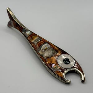 May include: A fish-shaped bottle opener with a metallic silver frame. The body is inlaid with iridescent mother-of-pearl and brown, swirled resin segments. A circular white and black eye detail is positioned near the open mouth of the fish.