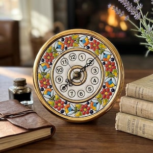 May include: A round ceramic clock with a gold-toned frame and a white face featuring black numerals from 1 to 12. The clock face is decorated with a colourful hand-painted floral border in shades of red, orange, yellow, green, and blue.