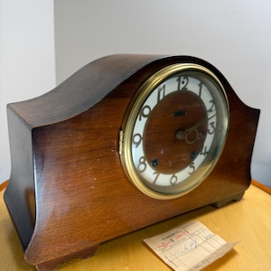 May include: A dark brown wooden mantel clock with a curved top and a circular face. The clock features a brass-coloured bezel, a white dial with black Arabic numerals, and two winding holes. A small paper tag with handwritten text sits nearby.