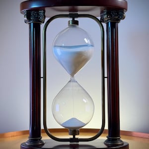 Peut inclure: Un sablier composé d'un bulbe en verre transparent rempli de sable blanc, maintenu par un cadre en métal doré. La structure est soutenue par deux colonnes en bois foncé avec des chapiteaux décoratifs, entre une base et un sommet ovales.