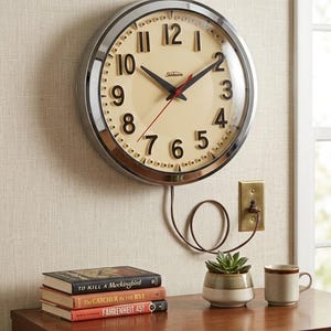 Peut inclure: Une horloge murale ronde Sunbeam avec un cadre chromé et un cadran crème doté de chiffres arabes noirs. L'horloge possède des aiguilles noires pour les heures et les minutes, une trotteuse rouge et un cordon d'alimentation marron branché.