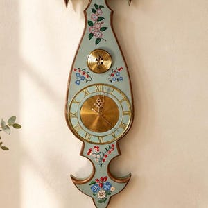 Peut inclure: Une horloge murale haute de couleur bleu clair avec un cadre en bois décoratif et des motifs floraux peints à la main en rouge, rose et bleu. L'horloge présente un cadran circulaire couleur laiton avec des chiffres romains et un baromètre.