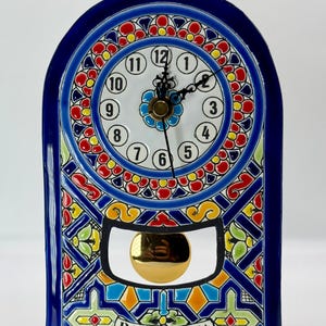 May include: An arched ceramic wall clock with a glossy deep blue border and a colourful hand-painted pattern in red, yellow, and green. The white clock face features black numerals from 1 to 12, black hands, and a gold-toned pendulum. The word BARCELONA is printed at the bottom.