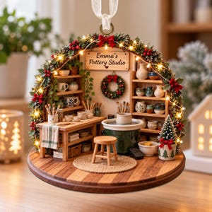 May include: A miniature pottery studio ornament featuring a wooden workbench, a green pottery wheel, shelves with ceramic mugs, and a small stool. The scene is framed by a festive evergreen garland with red berries and warm white fairy lights.