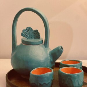 Può includere: Una teiera in ceramica color verde acqua con un manico a forma di foglia di ginkgo sul coperchio e un set di due tazzine abbinate. La teiera presenta un manico alto ad arco e un beccuccio corto. Le tazze hanno un interno arancione brillante.