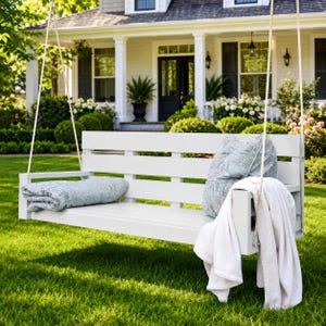 May include: A white wooden porch swing suspended by thick ropes, featuring a slatted backrest and flat armrests. The seat holds a folded blue and white patterned quilt, while a matching pillow and a draped white throw blanket rest on the side.