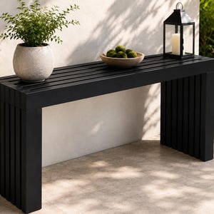 May include: A long, rectangular black bench featuring a slatted top and vertical slat legs. The bench is topped with a light-coloured stone planter containing a green plant, a wooden bowl filled with green moss spheres, and a black metal lantern.