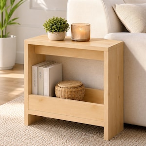 May include: A light-toned wooden side table featuring a flat top surface and a lower storage shelf. The table holds a small potted green plant, a ribbed glass candle holder, two books with visible spines, and a round woven lidded basket.