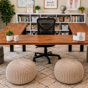 Puede incluir: Un escritorio de madera grande en forma de U con acabado de veta de madera marrón oscuro y patas de metal negro. El escritorio cuenta con una silla de oficina central de malla negra, dos pufs de punto beige y dos pufs verde salvia.