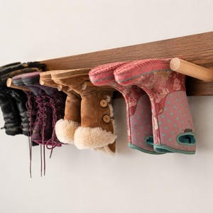 May include: A wall-mounted wooden boot rack featuring a dark brown horizontal plank with several light-colored cylindrical pegs. Four pairs of boots in various colors, including black, purple, tan with faux fur trim, and pink with teal dots, hang.