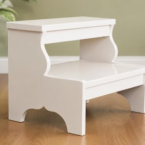 May include: A white wooden two-step stool featuring a smooth finish, a rectangular top step, a lower step, and curved side panels with arched cutouts at the base. The structure is solid and functional, providing two levels for reaching higher areas.