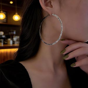 May include: A large, circular hoop earring in a polished silver tone. The surface of the hoop is textured with a dense, sparkling array of small, reflective crystals that catch the light, creating a continuous, shimmering effect along the entire ring.