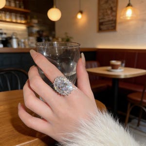 May include: A silver-toned ring featuring a large, oval-cut central stone surrounded by a halo of smaller, sparkling clear stones. The ring is worn on a finger, showcasing the reflective facets and brilliant light refraction of the jewellery piece.