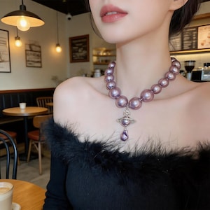 May include: A necklace featuring a strand of large, round, metallic purple pearls. A silver-toned pendant shaped like a planet with a ring around its center hangs from the strand, with a smaller, teardrop-shaped purple pearl dangling below it.