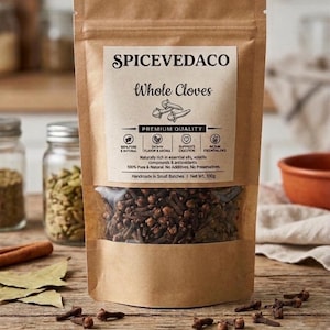 Puede incluir: Una bolsa de papel kraft marrón que contiene clavos de olor enteros con una ventana transparente. La etiqueta dice SPICEVEDACO Whole Cloves, Premium Quality, 100% Pure & Natural y Net Wt. 100g. La bolsa incluye iconos de sabor y digestión.