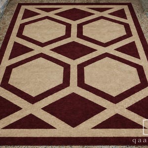 Pu&ograve; includere: Un tappeto rettangolare caratterizzato da un motivo geometrico di esagoni e rombi intrecciati color marrone scuro su uno sfondo beige chiaro. Il design include un bordo spesso e la scritta "qaartuz" nell'angolo in basso a destra.