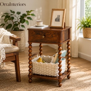 May include: A dark brown wooden side table with a single drawer and four spindle-style legs. The table features a lower shelf holding a white chunky knit basket with two handles, containing books and a pear. A picture frame sits on the tabletop.