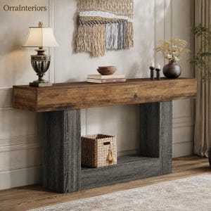 May include: A console table featuring a natural wood grain top and dark gray textured legs. The table includes a lower shelf holding a woven basket. On the surface sits a lamp with a cream shade, two stacked books, a bowl, and a dark vase.