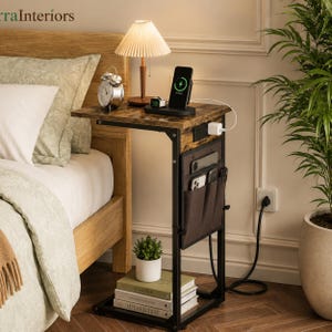 May include: A C-shaped side table with a dark wood grain top and black metal frame. It features a built-in power outlet, a side storage pocket, and a bottom shelf holding books and a small potted plant. A lamp, clock, and phone charger sit on top.
