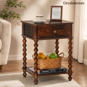 May include: A dark brown wooden side table with a single drawer featuring a round metal knob. The table has four turned spindle legs and a lower shelf. On top sits a white cup on a saucer and a picture frame. A woven basket with fruit sits below.