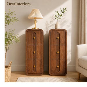 May include: Two tall, narrow wooden storage cabinets with rounded corners, each featuring three drawers with gold-toned circular knobs. The text OrraInteriors is visible in the top left corner. A pleated lamp sits on one cabinet and a vase on the other.