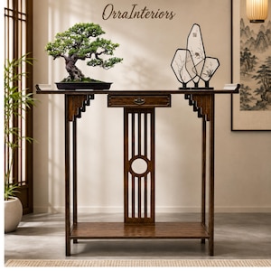 May include: A dark brown wooden console table with a single central drawer, a lower shelf, and vertical slat detailing featuring a circular cutout. On the table sits a green bonsai tree in a black pot and a black decorative frame with floral art.