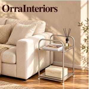 May include: A two-tier metal side table with a silver finish, featuring a top shelf holding a book and a reed diffuser, and a bottom shelf with a rectangular cream-colored box. The text OrraInteriors is visible at the top of the image.