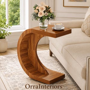 May include: A C-shaped side table crafted from medium-toned wood with a visible grain pattern. The table features a flat top surface and a curved base that extends to the floor. A glass vase with light-colored flowers and a white coffee cup sit on top.