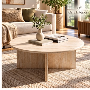 May include: A round, light-toned wooden coffee table with a smooth, circular top and a sturdy, cross-shaped base. On the table sits a small, cream-colored ceramic vase containing green leafy branches, alongside two stacked books.