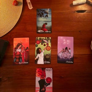 May include: A tarot card spread featuring five cards arranged in a cross pattern on a wooden surface. The cards display various illustrations including a person in a red robe, a person with a drum, a person holding a bird, and figures in white.