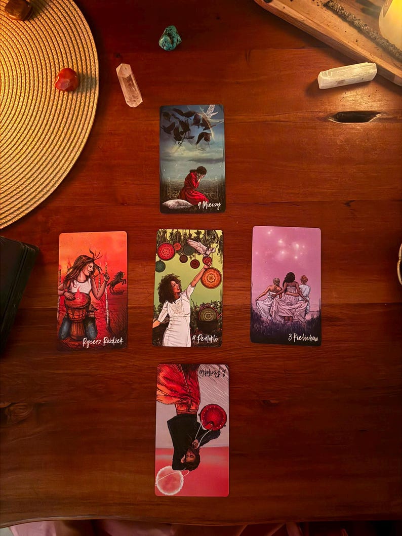 May include: A tarot card spread featuring five cards arranged in a cross pattern on a wooden surface. The cards display various illustrations including a person in a red robe, a person with a drum, a person holding a bird, and figures in white.
