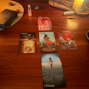 May include: A tarot card spread arranged in a cross pattern on a wooden surface, featuring five cards including the Eight of Pentacles, Five of Pentacles, and Page of Pentacles. The cards display colorful illustrations, and a deck of cards sits nearby.