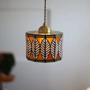 May include: A cylindrical stained glass pendant lamp featuring a repeating leaf pattern in white and amber tones. The glass panels are joined by dark metal soldering, and the fixture is suspended by a brown twisted cord from a brass-colored cap.