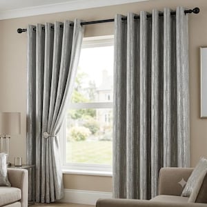 Op de afbeelding: Een paar vloerlange zilvergrijze gordijnen met een verticaal textuurpatroon en een metallic glans. De gordijnen hangen aan een zwarte metalen roede met ronde knoppen via zilverkleurige ringen, waarbij één paneel is vastgezet met een embrasse.