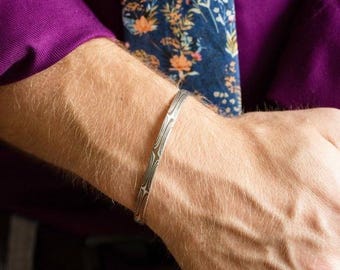 May include: A silver bracelet with a patterned design. The bracelet is worn on a person's wrist.