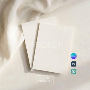 May include: Two stacked blank rectangular off-white cards with a textured paper finish. The cards are positioned against a soft, draped off-white fabric. The image includes the text MOCKUP and scene & studio in a thin white font.