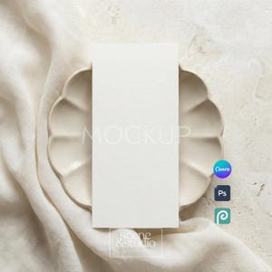 May include: A vertical rectangular white card mockup centred on a scalloped beige ceramic plate. The plate rests on a soft, draped off-white fabric. The card features the word MOCKUP in light grey text and the text scene & studio at the bottom.