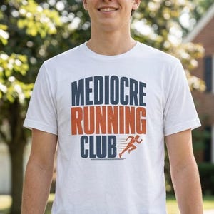 May include: A white short-sleeved t-shirt featuring the text MEDIOCRE RUNNING CLUB in dark blue and orange block letters, with a small orange graphic of a running person next to the word CLUB, worn with dark blue shorts and a coffee cup.