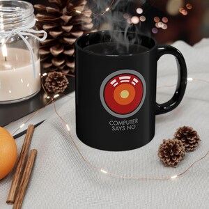 May include: A black ceramic mug featuring a circular graphic with a red outer ring, a yellow centre, and white horizontal bars. Below the graphic, the text "COMPUTER SAYS NO" is printed in white. The mug has a standard handle and a smooth finish.
