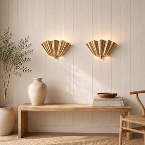 Peut inclure: Deux appliques murales dorées en forme d'éventail avec un design plissé rappelant un coquillage. Chaque applique présente une base centrale arrondie et diffuse une lueur chaude et douce, fixée sur un mur à panneaux verticaux au-dessus d'un banc.