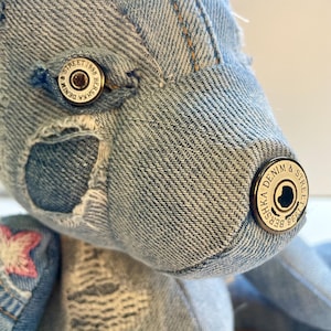 Può includere: Un orsacchiotto di peluche realizzato in tessuto denim azzurro con bordi sfilacciati e cuciture visibili. Gli occhi e il naso sono costituiti da bottoni metallici con impresso il testo "BERSHKA DENIM & STREET 1998". È visibile una piccola stella ricamata.